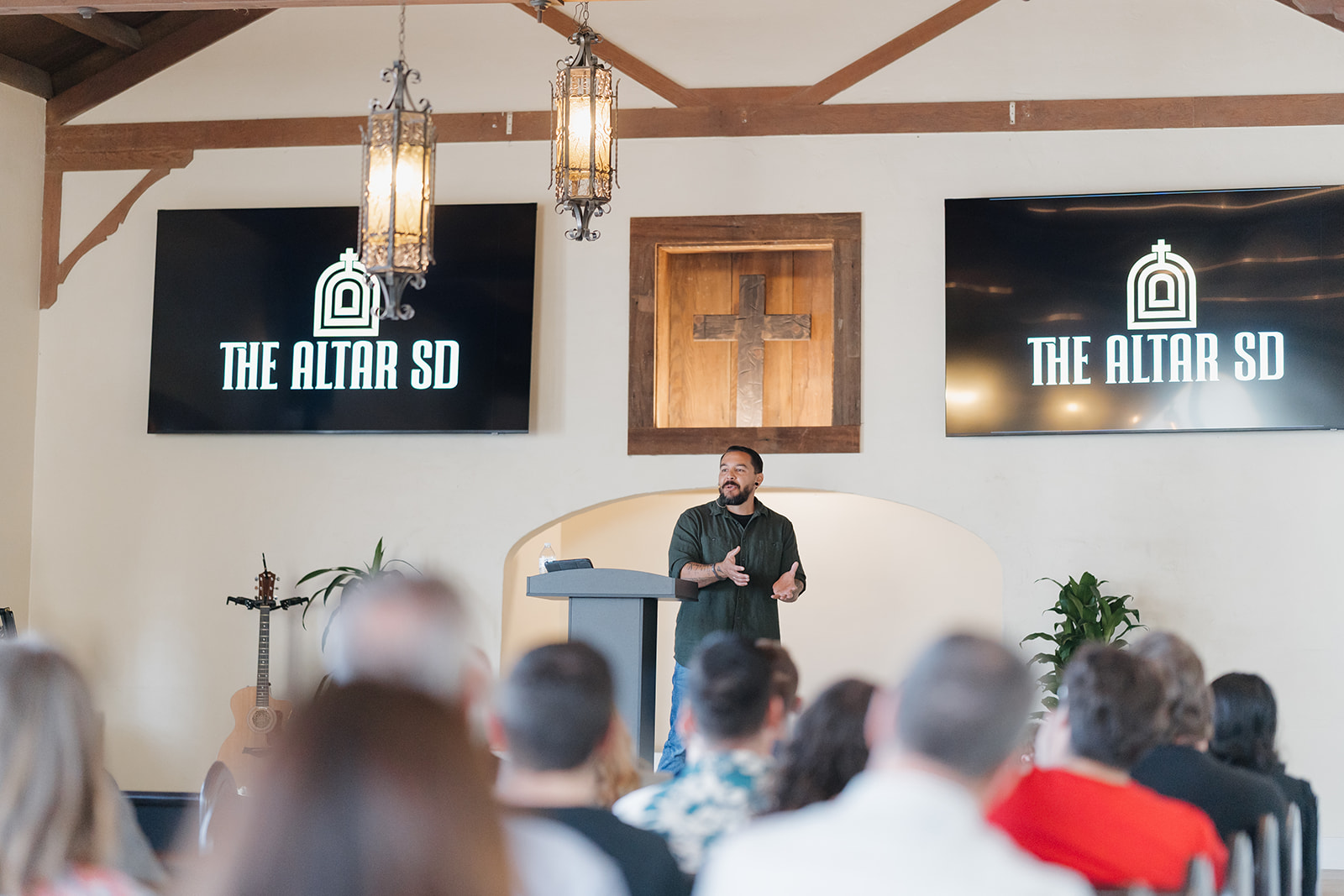 A speaker addresses the congregation at The Altar SD’s launch service in San Diego, with the church’s signs and a wooden cross behind him.